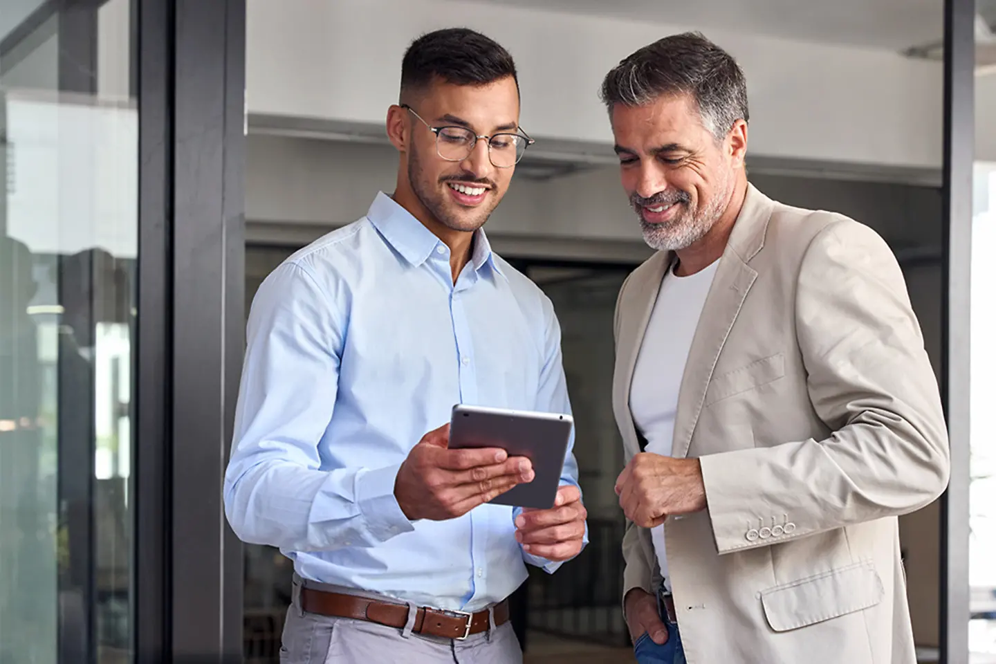 Deux hommes d’affaires de bonne humeur travaillent ensemble dans un bureau. Ils sont debout et utilisent une tablette numérique.