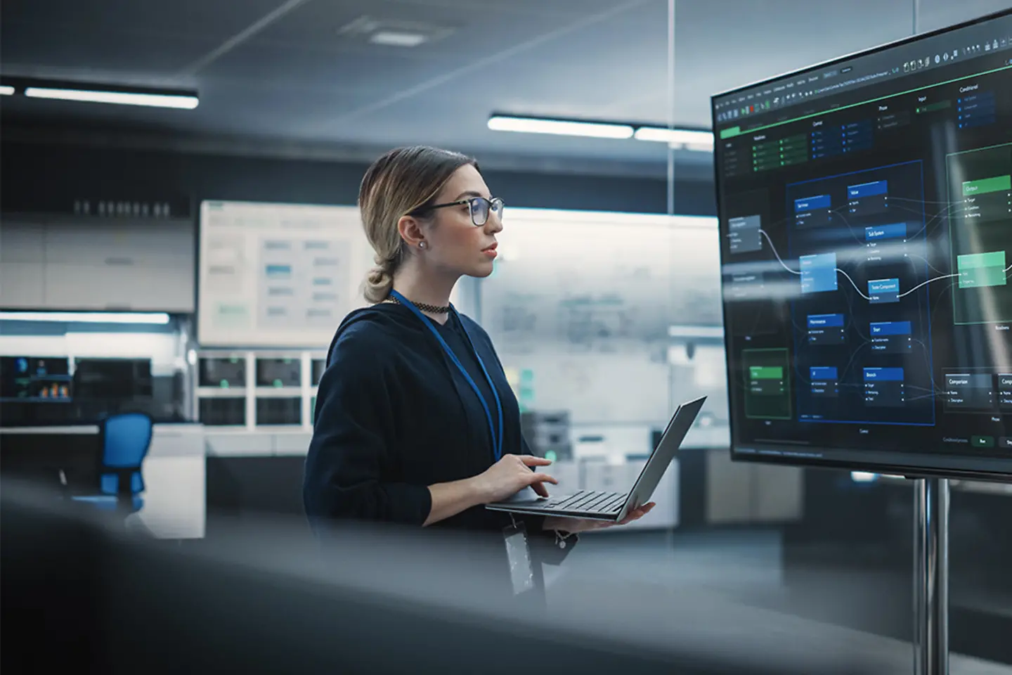 A female QA engineer works on a laptop computer while checking large display screens.